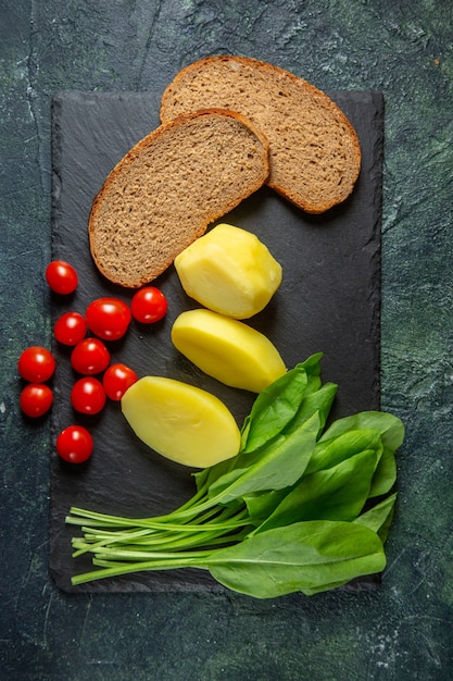 Vista verticale di patate fresche tagliate pelate e fette di pane dietetico pomodori fascio verde sul tagliere di legno sul verde nero mescolare i colori dello sfondo