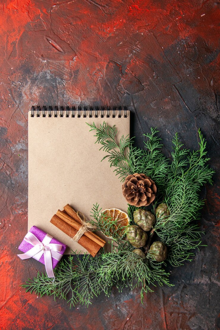 Foto gratuito vista verticale di rami di abete regalo di colore viola e taccuino a spirale chiuso lime di cannella su sfondo rosso