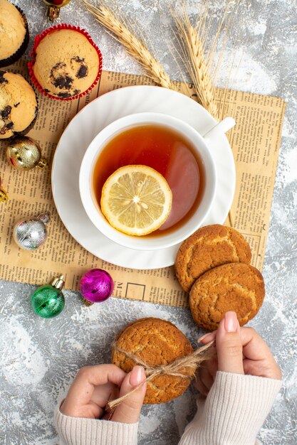 Vista verticale di una tazza di tè nero con limone e accessori decorativi su un vecchio biscotto di giornale e piccoli cupcakes sulla superficie del ghiaccio