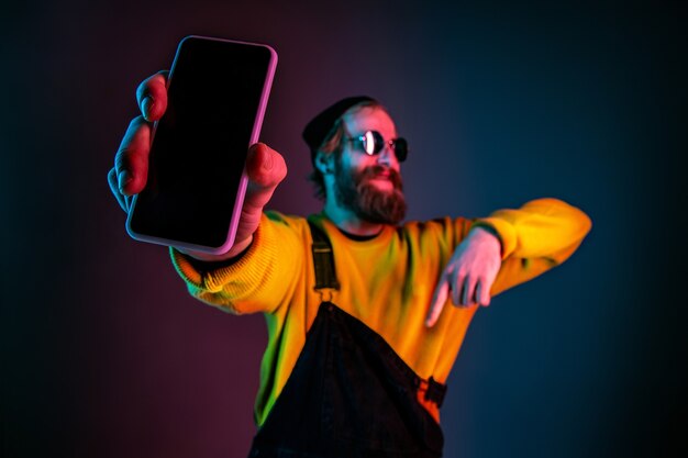 Visualizzazione dello schermo vuoto del telefono. Ritratto dell'uomo caucasico sul fondo dello studio sfumato in luce al neon. Bellissimo modello maschile con stile hipster. Concetto di emozioni umane, espressione facciale, vendite, annuncio.