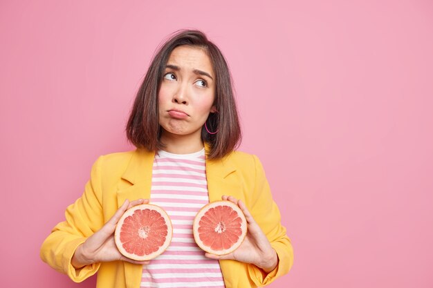 Vitamine fresche. La donna asiatica turbata tiene le metà del pompelmo ha pose di espressione triste con gustosa frutta succosa si sente scontenta continua a dieta vestita con abiti eleganti isolati su uno spazio vuoto muro rosa