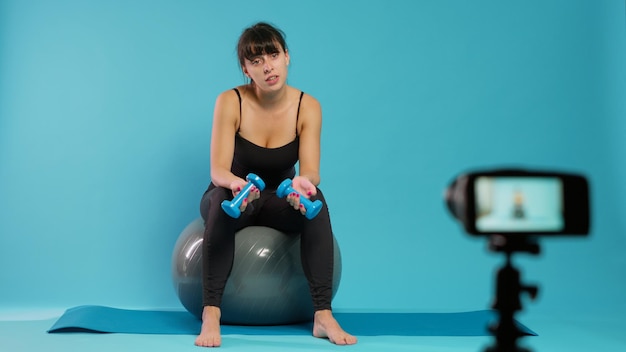 Vlogger di fitness che registra video di allenamento sulla fotocamera in studio, utilizzando manubri per spiegare la tecnica di sollevamento per la pratica sportiva. Donna muscolare che filma lezione di formazione online sulla palla tonificante.