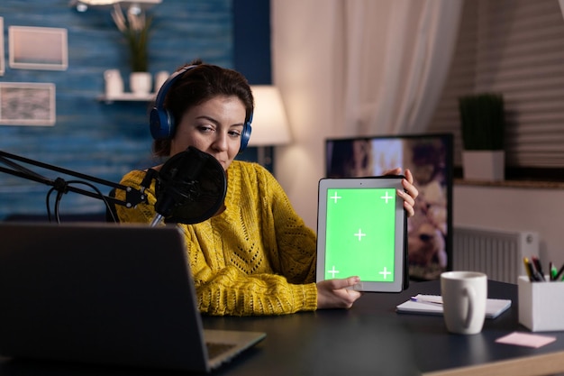 Vlogger donna con cuffie di produzione che tengono mock up tablet chroma key schermo verde con display isolato registrazione trasmissione online utilizzando apparecchiature podcast. Vlogger in streaming di contenuti live