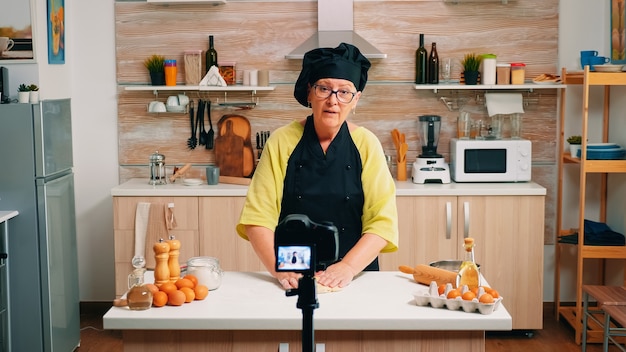 Vlogger femmina anziana che realizza video sui social media sulla cucina per il canale Internet. Blogger chef influencer in pensione che utilizza la tecnologia per comunicare, fotografare blog con apparecchiature digitali