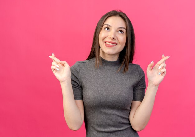 Volendo giovane donna graziosa che osserva in su mordere il labbro e facendo gesto di dita incrociate isolato su sfondo rosa con spazio di copia