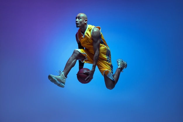 Volo. Bello giocatore di basket maschio afroamericano in movimento e azione alla luce al neon sulla parete blu.