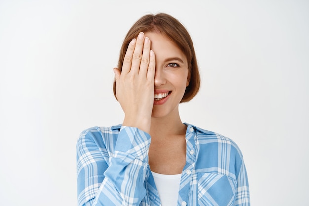 Volto di bellezza. Ritratto di giovane donna che copre metà del viso e sorridente, prima e dopo l'effetto cura della pelle, utilizzando crema per il viso o crema idratante, in piedi sul muro bianco