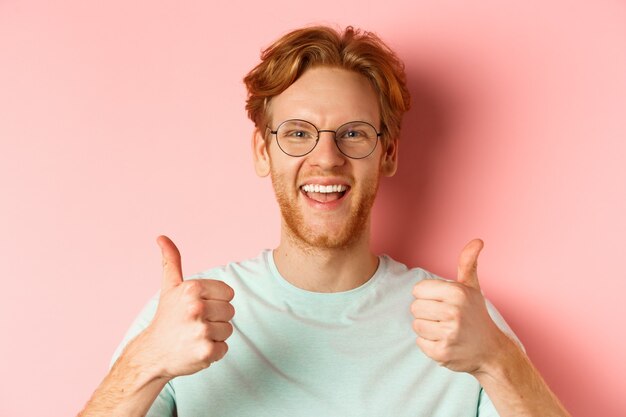 Volto di cliente maschio soddisfatto che mostra il pollice in su in approvazione, sorridendo felice, indossando occhiali e t-shirt, sfondo rosa.