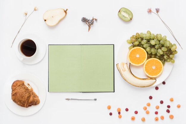 Vuoto notebook aperto con penna; croissant; caffè e frutta su sfondo bianco