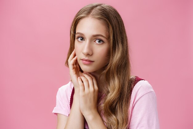 Woan si sente bella così com'è. Affascinante ragazza femminile e tenera con capelli naturali ondulati chiari e un leggero trucco che tocca delicatamente il viso prendendosi cura della pelle con cosmetici biologici.