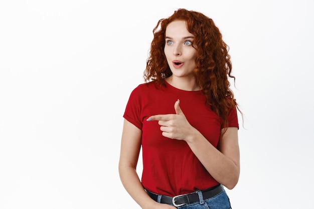 Wow dai un'occhiata a questa ragazza rossa impressionata con i capelli ricci che boccheggiano stupita indicando e guardando eccitata il logo sul lato sinistro che mostra lo sfondo bianco della pubblicità