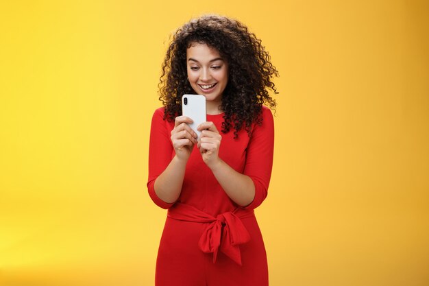 Wow nuovo telefono cellulare incredibile. Donna dai capelli ricci di bell'aspetto impressionata e stupita in abito rosso che tiene in mano uno smartphone che guarda lo schermo divertito mentre gioca a una bella app o a un gioco sul muro giallo.