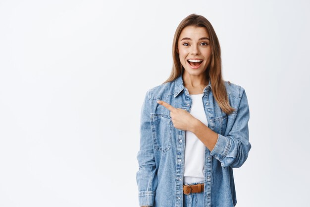 Wow, questo è buono, guarda qui. Sorridente ragazza carina con i capelli biondi che punta il dito a sinistra nello spazio della copia e fissa eccitata, mostrando una fantastica offerta promozionale, bianca