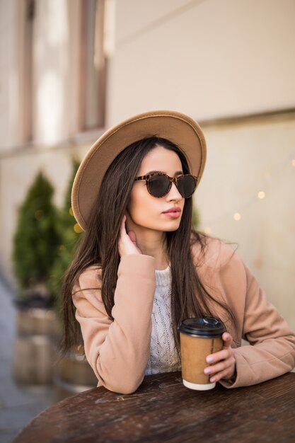 Yound fashion lady è seduta sul tavolo di abiti da caffè in abiti casual occhiali da sole scuri con una tazza di caffè