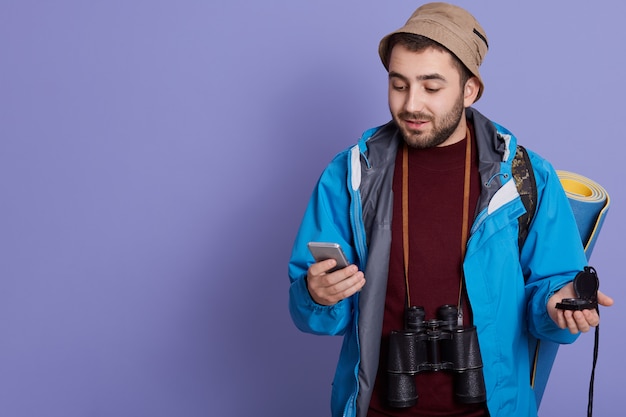 Zaino in spalla turistico uomo attraente utilizzando l'applicazione internet su smart phone moderno, in posa con tappetino, binocolo e zaino