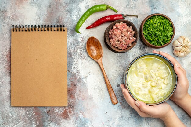 Zuppa di gnocchi di dushbara vista dall'alto in una ciotola in mano di donna aglio peperoncini piccanti cucchiaio di legno ciotole con carne e verdure un taccuino sulla superficie nuda