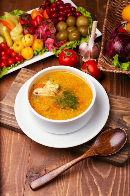 Zuppa di pollo con un pezzo di carne in salsa di pomodoro. Prezzemolo tritato, in una ciotola bianca decorata con turshu sul tavolo di legno.