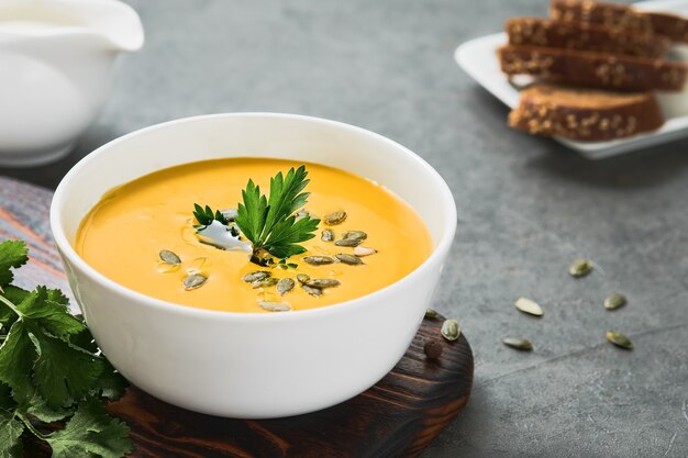 Zuppa di zucca e carote con olio d'oliva in una tazza bianca, semi di zucca e prezzemolo su sfondo grigio. Primo piano su zuppa vegetariana di stagione, affilatura selettiva su prezzemolo