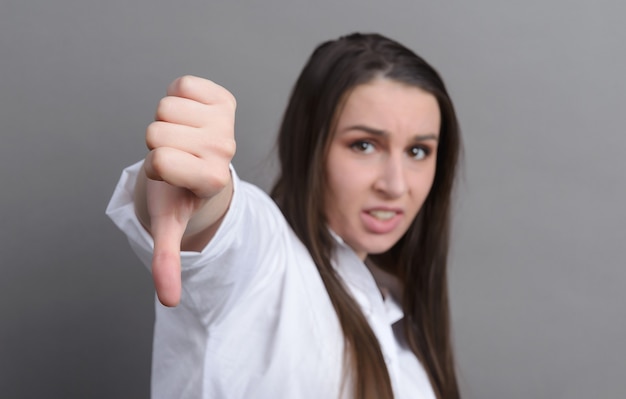 1 giovane donna bianca con una camicia bianca mostra un segno con il dito verso il basso, cattivo su una superficie grigia