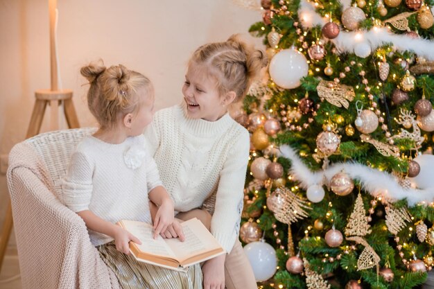 2 ragazze bionde sono sedute su una poltrona vicino all'albero di Natale e leggono un libro serate accoglienti con un libro vicino all'albero Le amiche leggono un libro insieme Relazione calorosa