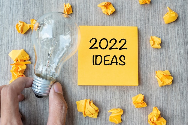 2022 parole di idea sulla nota gialla e carta sbriciolata con l'uomo d'affari che tiene la lampadina sul fondo della tavola di legno. Nuovo anno nuovo inizio concetto creativo, innovazione, immaginazione, risoluzione e obiettivo