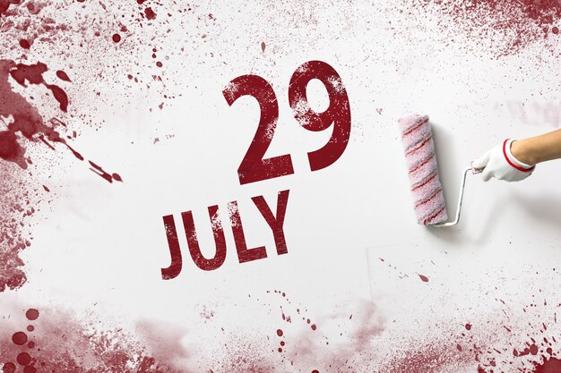 29 luglio. Giorno 29 del mese, data del calendario. La mano tiene un rullo con vernice rossa e scrive una data di calendario su uno sfondo bianco. Mese estivo, concetto di giorno dell'anno.