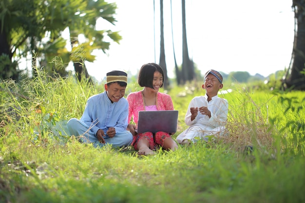 3 bambini in abiti musulmani si siedono e guardano Internet sul portatile per divertirsi nel giardino agricolo.