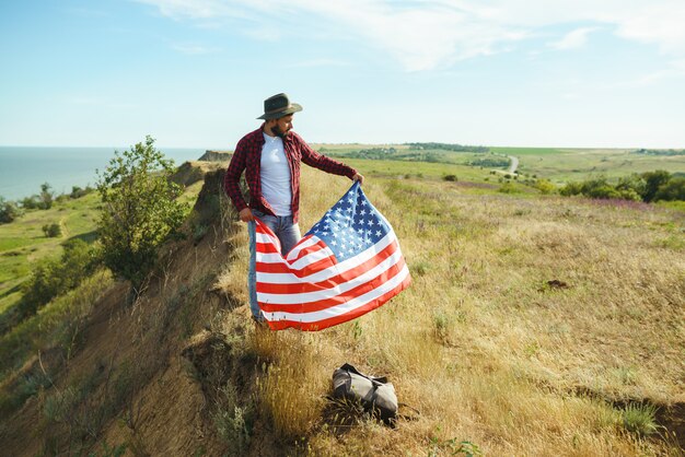 4 luglio. Quattro luglio. Americano con la bandiera nazionale. Bandiera americana. Giorno dell'Indipendenza. Vacanze patriottiche. L'uomo indossa un cappello, uno zaino, una camicia e jeans.