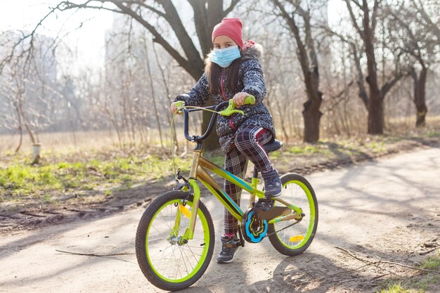 7 anni felice Bambina che va in bicicletta nel parco a casa e indossa una maschera di protezione per proteggere pm2.5 e sintomi del virus pandemico Coronavirus Covid-19. Esercizio sportivo per la salute.