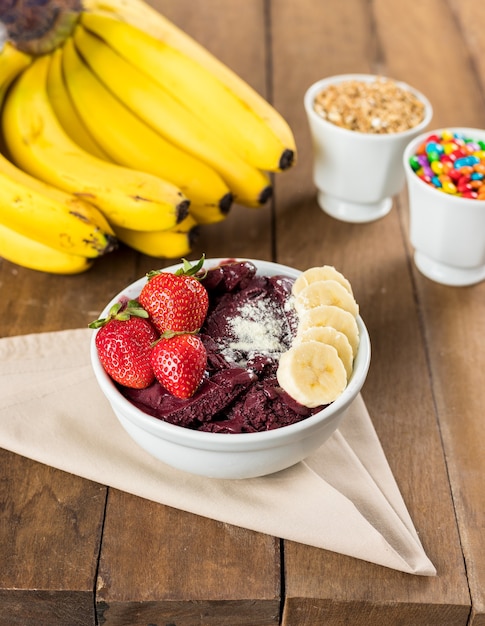 AÃ§ai brasiliano su una pentola bianca con bacche di paglia, banana e latte in polvere su uno sfondo di legno e frutta in estate.