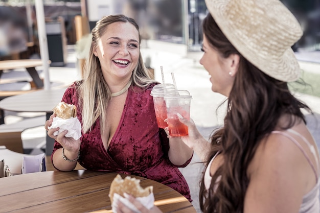 A noi. Donne allegre felici che tifano con bicchieri di limonata mentre mangiano hamburger nella caffetteria