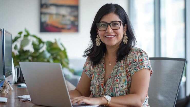 a woman in glasses is sitting on a laptop and smiling