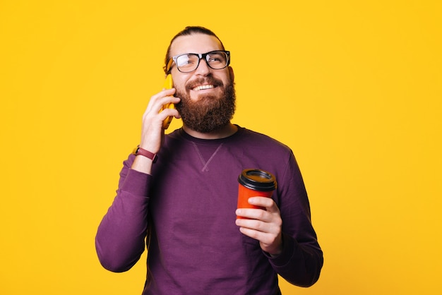 A young bearded man is talking to his phone smiling is holding a cup of hot drink