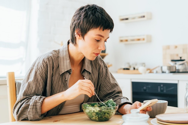 A young short-haired brunette  in the kitchen eating and browsing social networks on her phone