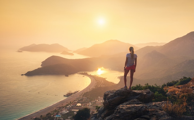 Abbellisca con la ragazza, il mare, le creste della montagna e il cielo arancio