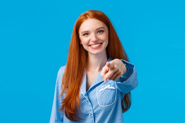 Abbiamo bisogno di voi. Ragazza rossa allegra e fiduciosa facendo una scelta sicura, indicando il dito e sorridendo come decide, scegliendo tra le varianti, parlando con una persona, in piedi nella parete blu del pigiama