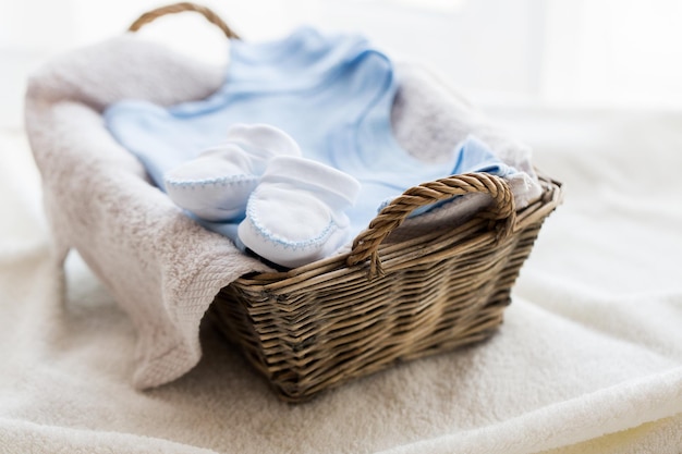 abbigliamento, infanzia, maternità e concetto di oggetto - primo piano di stivaletti bianchi con mucchio di vestiti e asciugamano per neonato nel cesto sul tavolo
