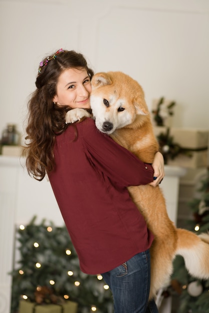 Abbracci di bella donna, coccole con il suo cane Akita Inu. su uno sfondo di un comò di albero di Natale con candele in una stanza decorata. felice anno nuovo e buon Natale