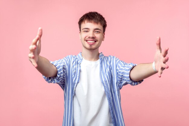 Abbracci gratis. Vieni qui. Ritratto di uomo dai capelli castani gentile e amichevole con piccola barba e baffi in camicia a righe casual che allunga le mani per abbracciare. studio indoor girato isolato su sfondo rosa