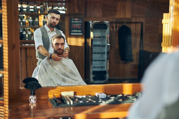 Abile barbiere maschio che fissa un mantello intorno al collo del suo cliente prima di mettersi a lavorare sui suoi capelli