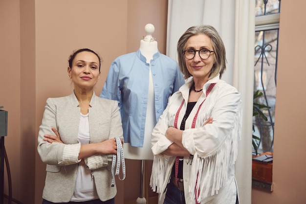 Abile e sicuro team di due sarti donne che posano con un manichino manichino che indossa una camicia blu della nuova collezione in un atelier di sartoria