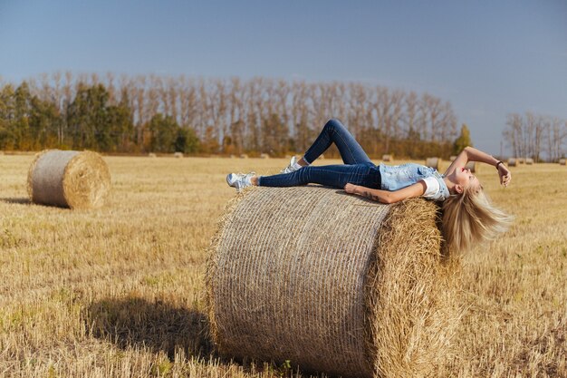 Abitante del villaggio della bella giovane donna che posa in jeans su una balla di fieno in un campo