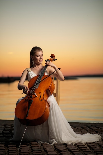 Abito bianco da ragazza che suona il violoncello sulla riva del lago dopo il tramonto, perfetto per una copertina o un poster