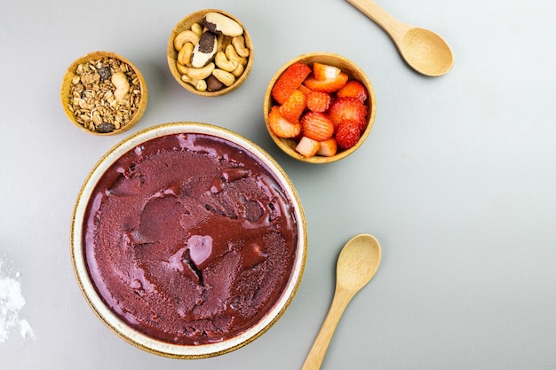 Acai brasiliano in una ciotola bianca granola alla fragola aggiuntiva e noci in piccole ciotole di bambù Anacardi e Para Vista dall'alto Sfondo grigio Messa a fuoco selettiva