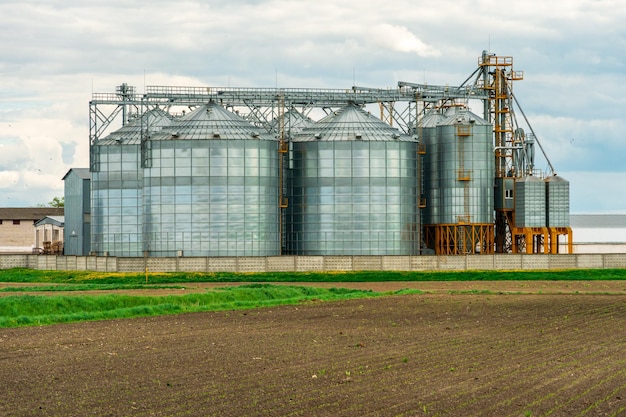 Accanto al campo agricolo arato installato silos d'argento su impianto di produzione agro per la lavorazione di essiccamento pulitura e stoccaggio di prodotti agricoli farina cereali e grano Ascensore per granaio