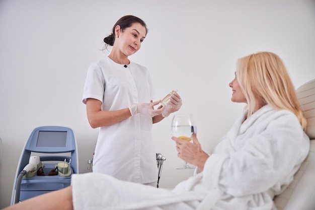 Accappatoio bianco donna bionda felice che tiene un bicchiere d'acqua mentre ascolta il suo cosmetologo nella stanza al chiuso