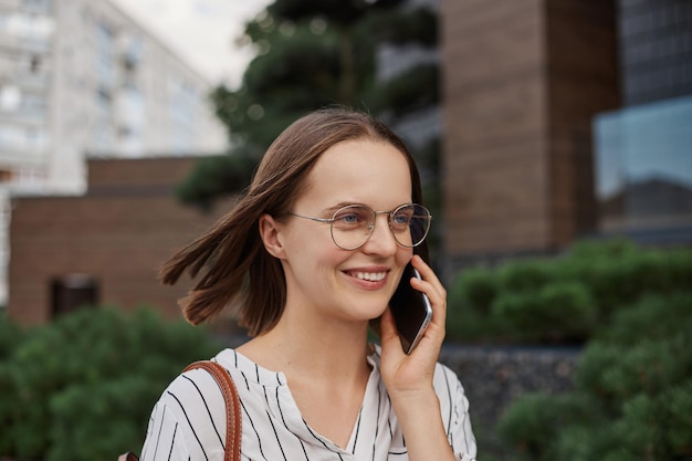 Accattivante giovane donna dai capelli castani che indossa camicetta che parla al telefono cellulare distogliendo lo sguardo camminando per la strada della città esprimendo emozioni positive