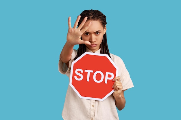 Accesso negato Donna prepotente con i dreadlocks che mostra il gesto di arresto con la mano e tiene il cartello rosso del segnale di stop entrare è limitato indossando una camicia bianca Scatto in studio al coperto isolato su sfondo blu