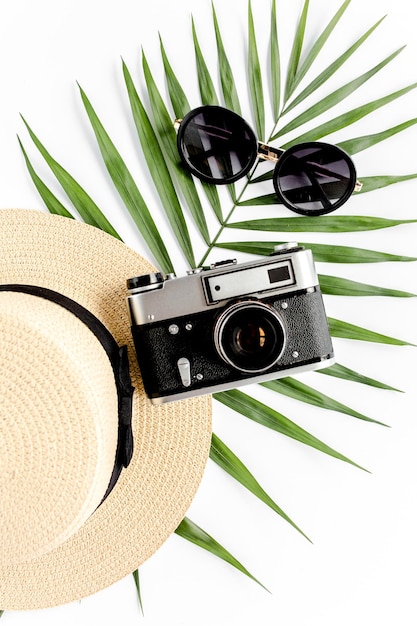 Accessori da viaggio piatti su sfondo bianco con occhiali da sole con cappello di paglia per fotocamera retrò e foglia di palma tropicale Sfondo del concetto di viaggio