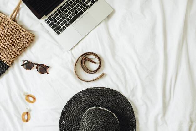 Accessori moda donna, occhiali, orecchini, cintura, cappello di paglia, borsa di paglia e laptop sdraiato sul letto con lenzuola bianche